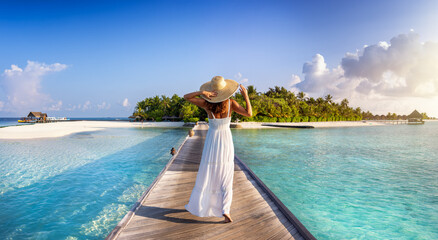 Back view of a elegant woman in white dress and hat walks down a pier towards a tropical island in the Maldives - 637516233