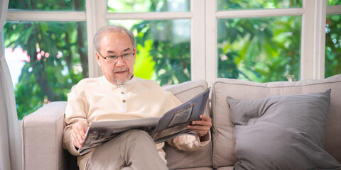 Senior asian retirement old man in casual outfit wearing glasses sitting and reading newspaper in modern house, Asian senior man sitting on sofa and reading newspaper at home.