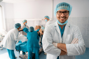 Portrait of a handsome smiling successful medical professional wearing eyeglasses and surgical cap with arms crossed, standing in delivery room in front of medical team and woman in labor. - obrazy, fototapety, plakaty