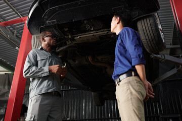 Car owner and mechanic standing under suspended car and discussing what needs to be fixed
