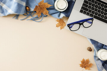 Desk adorned in fall aesthetics. Top view shot of candles, eyewear, warm blanket, laptop, acorns, dry autumn leaves on pastel beige background with blank space for promo or message