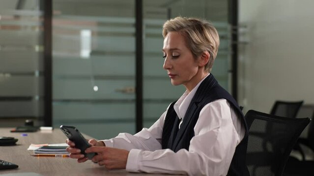 Side view of serious middle-aged businesswoman using smartphone sitting at office desk touching screen, typing message on mobile phone, browsing Internet, social media, shopping online, using app.