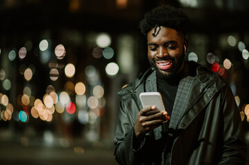A young happy African American man is standing on the street at night with earbuds on and smiling...