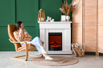 Young woman with cup of coffee talking by mobile phone in armchair near fireplace at home