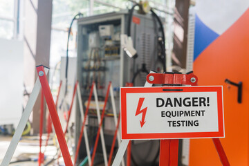 sign with the inscription Danger! Equipment testing! on the fence against the background of blurred electrical facilities in the workshop