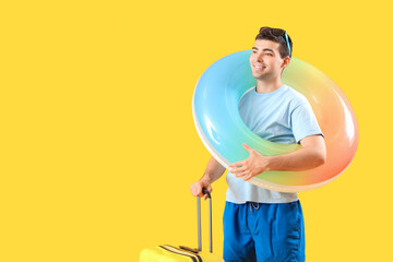 Male tourist with suitcase and swimming ring on yellow background