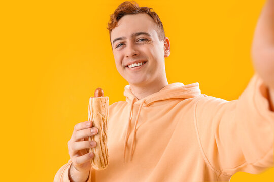 Young man with tasty hot dog taking selfie on yellow background