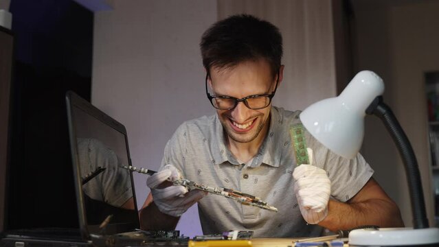 Portrait of happy laptop repairman with glasses. Middle aged man examines motherboard of disassembled laptop, finds cause of breakdown. Engineer smiles, rather shakes circuit board in gloved hand.