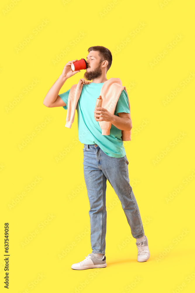 Wall mural Portrait of handsome young man with tasty hot dog drinking coffee on yellow background