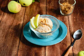 Two scoops of vanilla ice cream with shortbread crumble and caramelized apple slices in a transparent bowl on a brown wooden background.