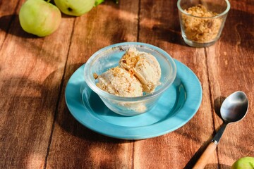 Two scoops of vanilla ice cream with shortbread crumble and caramelized apple slices in a transparent bowl on a brown wooden background.