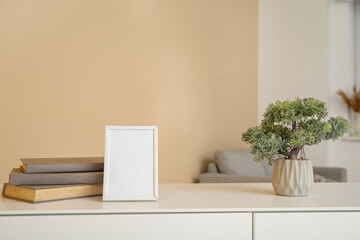 Blank frame, decorative tree in pot and books on white cabinet near beige wall