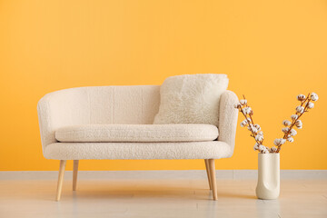 White sofa with cushion and cotton branches in vase near orange wall
