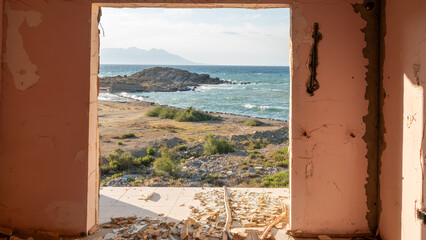 View of coastline through the broken window in a room of abandoned and demolished old hotel building. View through a broken window to the sea. 