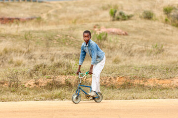 african child in the village riding a small bike on a dirt road going to school