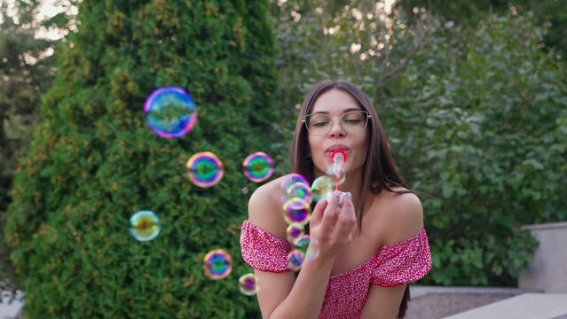 Beautiful Happy Woman Blowing Soap Bubbles on Street. Childhood in Adulthood. Smiling Face Optimistic Girl in Glasses Having Fun with Child toys. Funny Woman Playing Children's Game. Slow Motion.