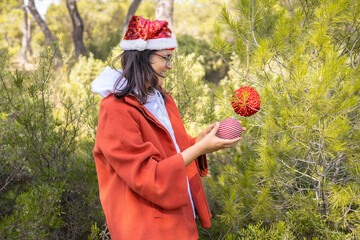 Caucasian middle age woman in her 40s with an orange coat and a christmas hat decorated with sequins, setting the christmas tree outdoors in a sunny day. Normal people.