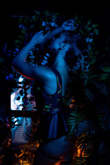 Fototapeta na wymiar Portrait of a beautiful go-go dancer posing in a nightclub against the backdrop of plants. Blue atmospheric lighting. Party concept, nightclub