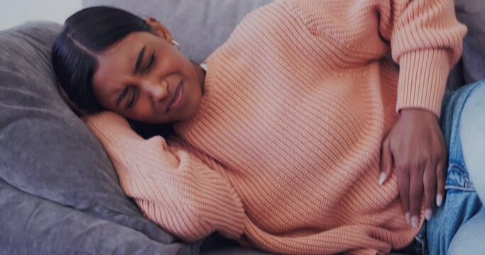 Woman, sick and period pain on home sofa with endometriosis, colon crisis and ibs constipation. Indian person, stomach ache and stress from tummy virus, gut health risk and gastrointestinal bloating