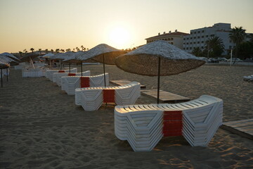 Gestapelte weiße Ligen aus Kunststoff und Sonnenschirme aus Korbgeflecht im warmen Licht der...