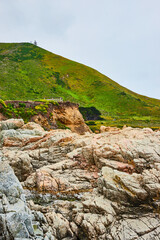 Baren rock cliff face with jagged crags and distant green mountainous hillside under overcast sky