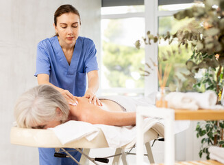 Young female masseuse doing back massage to elderly woman client in massage room