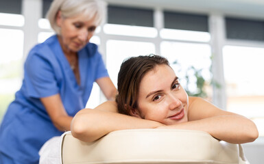 Positive young adult woman relaxing in cozy spa center while professional masseuse doing back...