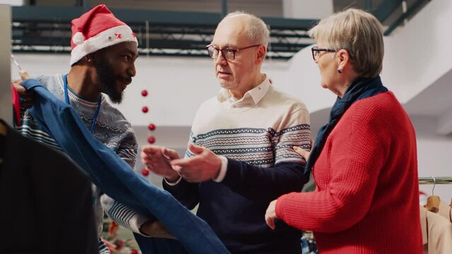 Retail assistant in xmas adorn fashion boutique helping old couple during their Christmas frenzy shopping spree. Worker offering advice to aged clients during festive holiday season