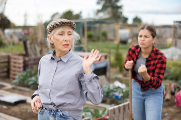 Two disappointed young and old female neighbors quarreling while digging garden during daytime in...