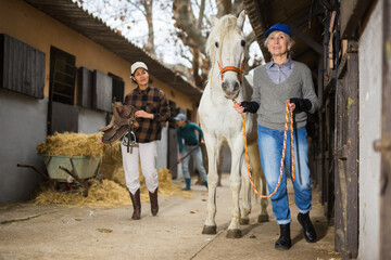 Women ranchers preparing white horse for ride. Senior European woman leading horse, Asian woman carrying saddle.