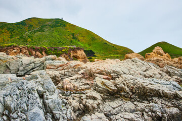 Baren rock cliff face in jagged crags with distant green mountainous hillside and overcast sky