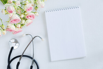 Bouquet of pink rose flowers and stethoscope with empty card notebook on a white background. National Doctor s day greeting card. Top view flat lay, copy space