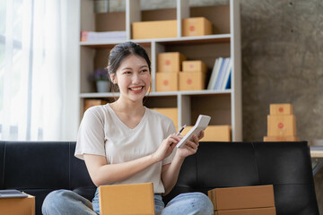 Pretty smiling Asian woman online shopping at home with parcel boxes, online delivery, small SME business owner. Seller preparing parcel boxes to send to customers. Online selling or e-commerce