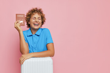 Smiling girl with short curly hair dreaming about future voyage, dressed blue shirt, holding passport with smile and closed eyes on pink background, tourism concept, copy space
