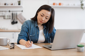 Woman making notes of article from computer in apartment