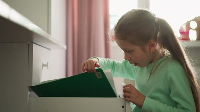 Little girl opens drawer and wonders of found folder in children room. Student seeks craft items in commode container at home. Schoolgirl and furniture