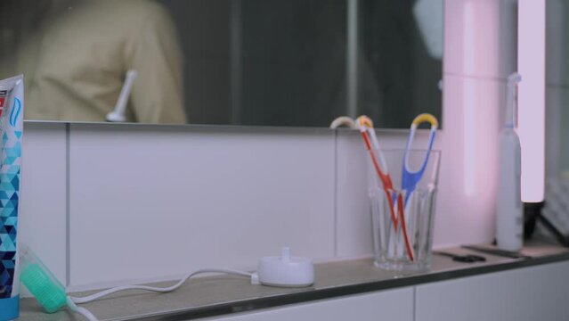 Man removes electric toothbrush from charge and brushes teeth before work in morning. Male in shirt picks up the ultrasonic dental brush view of reflection in mirror. Dental cleaner power tool. 