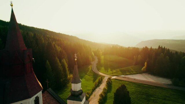 The drone flies past a monastery with a chapel standing among a dense spruce forest on top of a mountain. Drone video at sunset of a summer day