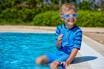 Smiling blond boy in a blue bathing suit sits on the side of a summer pool, his legs lowered into...