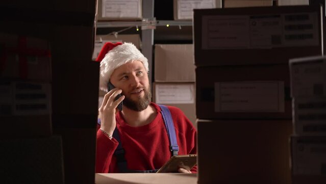 Handsome warehouse worker wearing santa hat uses digital tablet for checking stock, he is calling at cellphone near shelves with cardboard boxes.