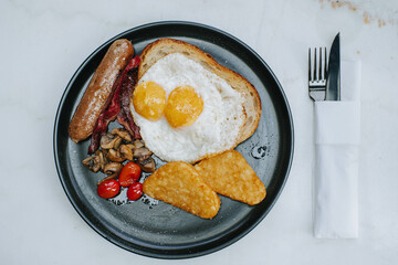Flat lay or top view shot of a plate of Full English Fry Up Breakfast contains hashbrown with sunny side egg, sourdough, bacon, sausage, mushrooms, and cherry tomatoes with knife and fork on the right