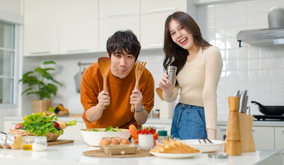 Obraz na płótnie Canvas Asian young lovely lover couple husband and wife in casual outfit standing smiling holding red tomato while mixing vegetable salad with wooden fork and spoon in bowl in full decorated modern kitchen