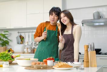Obraz na płótnie Canvas Asian young lover couple husband and wife in casual outfit with apron standing smiling posing holding wooden spoon and fork utensil in full decorated modern kitchen with cooking equipment at home