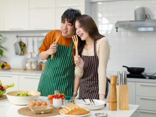 Asian young lover couple husband and wife in casual outfit with apron standing smiling posing holding wooden spoon and fork utensil in full decorated modern kitchen with cooking equipment at home