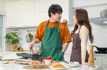 Asian young lover couple husband and wife in casual outfit with apron standing smiling while man cooking frying food with pan woman helping in full decorated modern kitchen with ingredients equipment