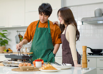 Obraz na płótnie Canvas Asian young lover couple husband and wife in casual outfit with apron standing smiling while man cooking frying food with pan woman helping in full decorated modern kitchen with ingredients equipment
