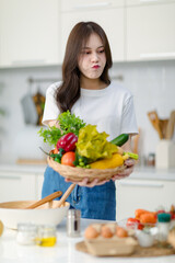 Obraz na płótnie Canvas Asian young beautiful housewife in casual outfit standing smiling showing red sweet pepper and mixed fresh organic vegetables basket in full decorated modern kitchen with ingredients and equipment