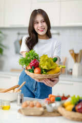 Obraz na płótnie Canvas Asian young beautiful housewife in casual outfit standing smiling showing red sweet pepper and mixed fresh organic vegetables basket in full decorated modern kitchen with ingredients and equipment