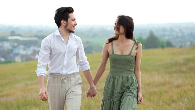 Happy couple of young man and woman smiles walking by hand across field on hilly plain. Lovers joy and landscape against city slow motion