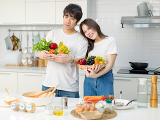 Obraz na płótnie Canvas Asian young lovely lover couple husband and housewife in casual outfit standing smiling posing holding mixed fruits and vegetables basket in full decorated modern kitchen with ingredients equipment